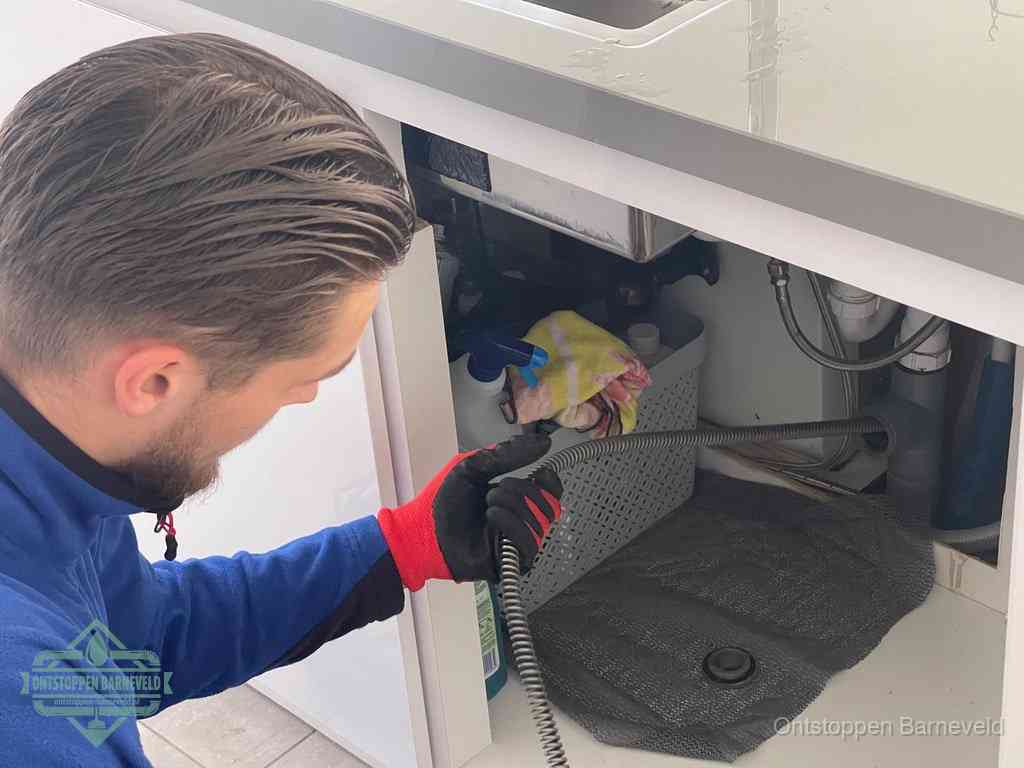 Een monteur voert een flexibele ontstoppingsveer in onder een keukenkastje om een verstopping voorbij de sifon te bereiken in Barneveld