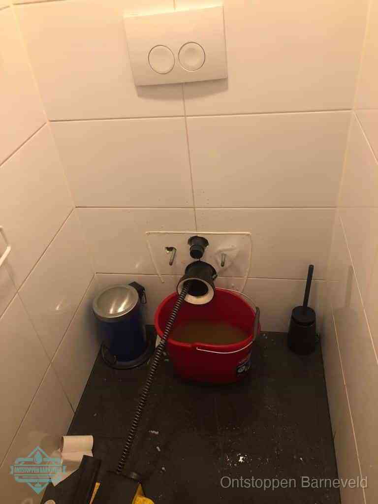 Een ontstopte wand-wc met verwijderde toiletpot waar een ontstoppingsveer in de wanduitgang verdwijnt, het achterhalen waar de verstopping zit in Barneveld