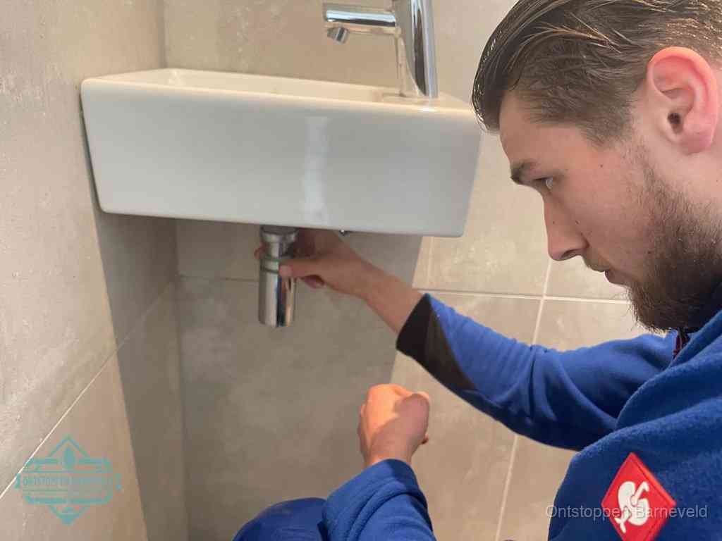 Een monteur draait met de hand de zwanenhalssifon onder een fonteinwasbak in de toiletruimte los om de bron van rioollucht in Barneveld te vinden